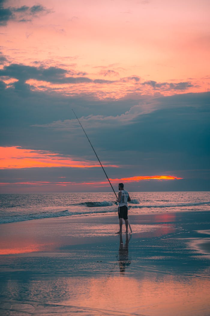 A solitary man fishing on a serene beach at sunset, with beautiful pastel skies reflected in the water.